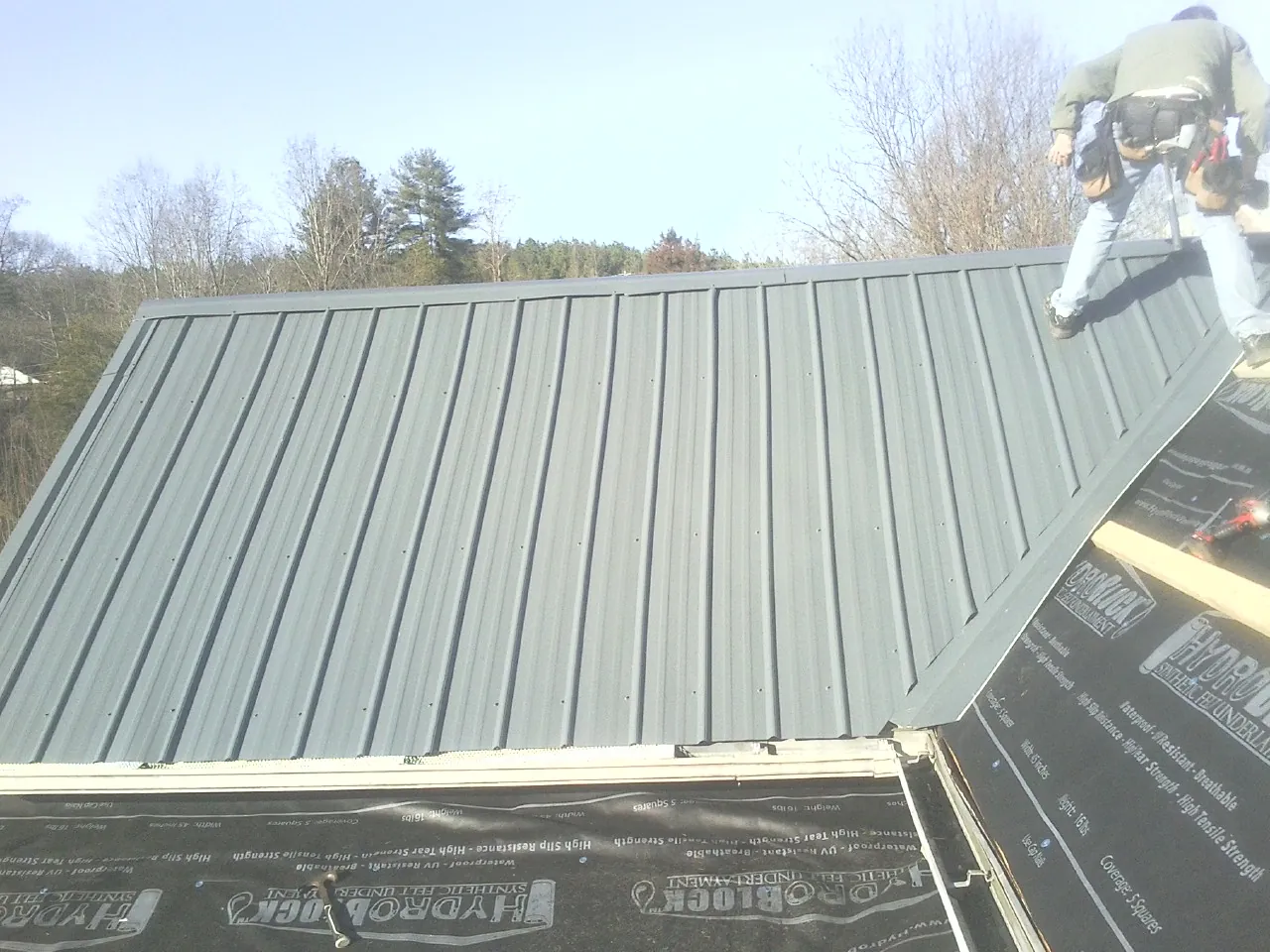 Crew installing dark green standing seam panels over synthetic underlayment in Burton