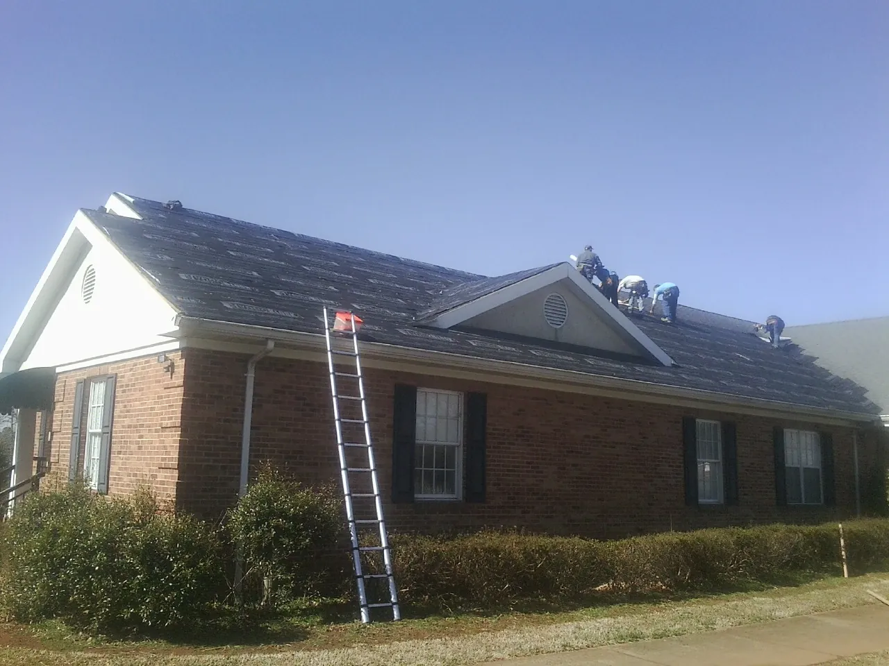 Crew tearing shingles off a brick ranch roof with a ladder set — Metal Roof Repair work by our crew in Burton