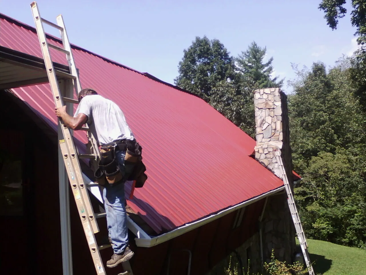 Roofer on a ladder beside a red metal roof and stone chimney — Soffit & Fascia Repair by Roofing Brothers in Burton